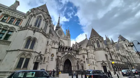 Stuart Woodward/BBC The Royal Courts of Justice building in London. It is a grey concrete building with ornate detailing, turreted roof and archways. There is road traffic in front of the building and blue sky and grey clouds above