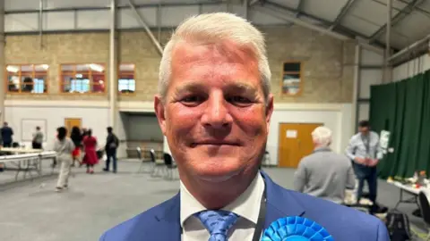 Stuart Andrew with short white hair wearing a blue suit and blue rosette