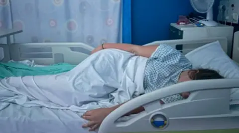 Grace A young woman is lying in a hospital bed wearing a hospital gown and partly covered by a white sheet. We can't see her face as it is hidden by the bed arm. She seems like she may be asleep.