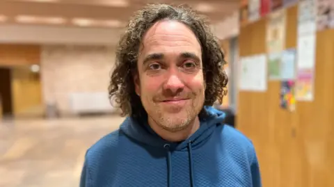David Russell looking into camera in a community centre. He has shoulder length dark curly hair, stubble and is wearing a blue hooded jumper.