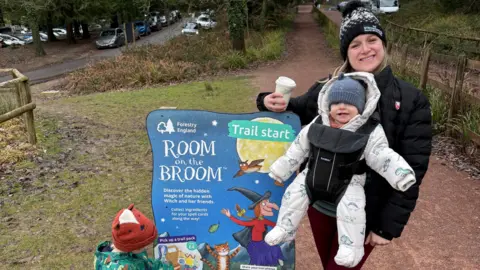 Tzara Spurrier A woman wearing a black coat and bobble hat, with a babyin a sling on her front, standing next to a forestry england trail sign
