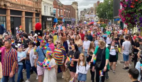 A large group of people making their way along Lincoln High Street. Some are brightly dressed in rainbow colours and some are holding flags. Other people are standing watching the parade.