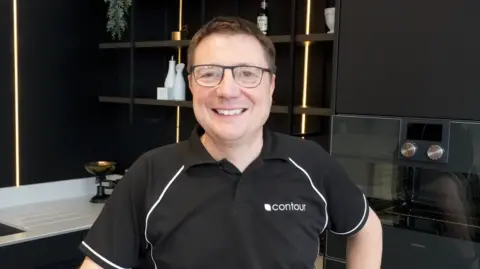 David Burgess is smiling at the camera. He has glasses with black rims and is wearing a black polo shirt with writing on the left side which reads 'Contour'.