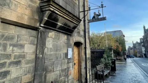 Photo of the building in Edinburgh's tollbooth. It is the side of a blonde sandstone building. It looks old and has a wooden door with iron rivets