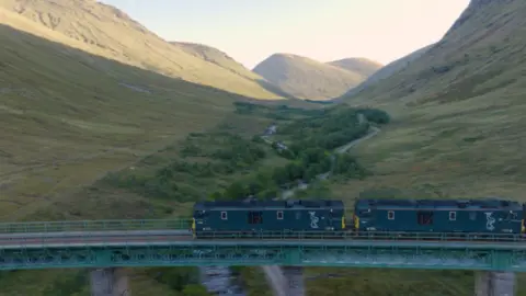 A green locomotive crosses a bridge in the Scottish Highlands