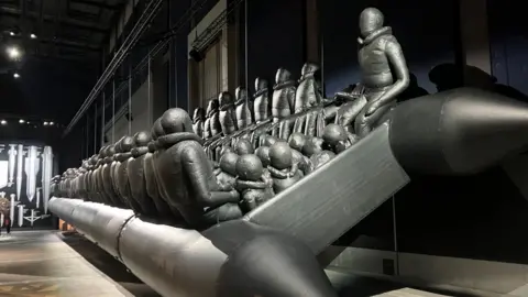 Photograph of the Ai Weiwei: Button Up! exhibition at Manchester's Aviva studios. The image shows a giant black inflatable dinghy with dozens of dummy refugees.