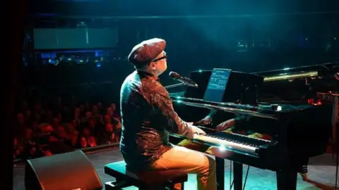 Contributed Phil Jackson during a concert. He is playing a piano on a stage in front of a large crowd. He wears a flat cap, shirt and cream trousers. 