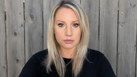 A woman with blonde hair in a black jumper and serious facial expression looking at the camera. She is standing in front of a grey garden fence.