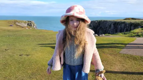 Family photo Gwen Harries looking at the camera. She is stood on grass at the edge of a cliff with the sea behind her. She is wearing a pink coat and a pink and white spotty hat.