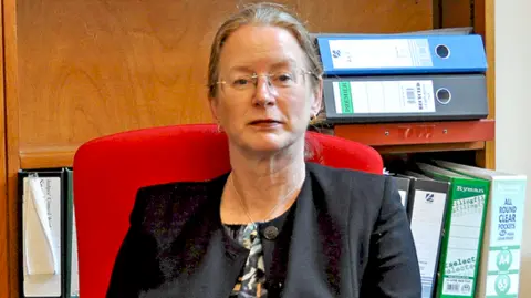 Office of the Chief Coroner Alexia Durran poses seated for a photograph in her office with large files on shelves behind her. She has glasses and tied-back, fair hair and wears a black jacket over a multi-coloured blouse.