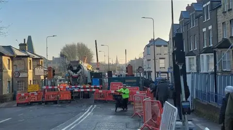 The Botley Road closure from the side leading to the city centre. There are multiple barriers surrounding the pavement and the road. A lorry can be seen behind them. A person with a helmet and a yellow high vis jacket can be seen pushing a bike on the road. There are people walking on the pavement. The city spires and Said Business School can be seen in the distance