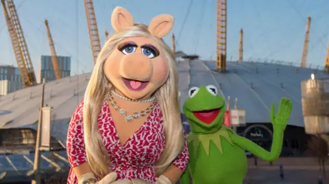 Getty Images Miss Piggy wears a white and pink dress and silver jewellery. She stands beside Kermit the Frog who is waving. The pair are standing in front of The O2 in London.
