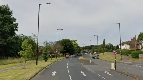 A road with two lanes and a filter lane turning right, with parked cars in the distance. The road is lined with grass verges and trees and there are lampposts uniformly spread out along both sides.