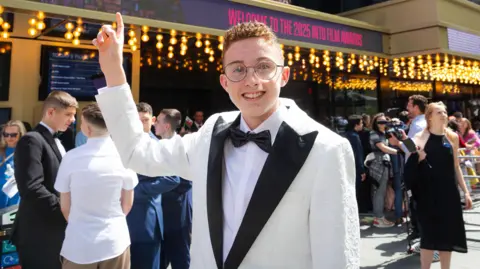 A young man with red, short curly hair and classes points upwards while wearing a white suit with black lapels and a black bow tie with a crowd milling about in the background