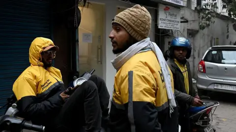 Getty Images Three men, wearing yellow-and-black windcheaters with the logo of a quick delivery company, stand near their two-wheelers, in Delhi's Janpath during a nationwide strike of gig workers on 31 December 2025