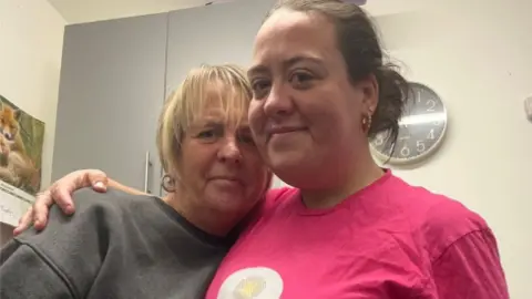 Two women stand close together, one has an arm around the other. The woman on the left has bobbed blonde hair, the woman on the right has dark hair