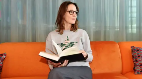 A woman in a grey top is sat on an orange sofa, and is holding an open book. She is looking to the side of the camera
