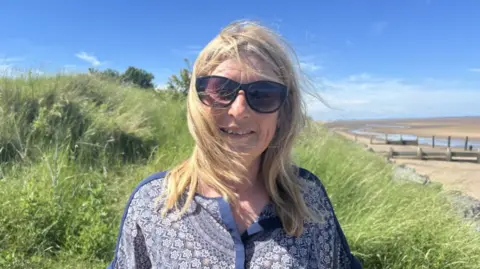 Caroline Carr - a woman with long blonde hair, wearing sunglasses and a blue and white patterned dress. She is standing near long grass and next to a sandy beach with blue sunny skies.