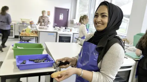 Roger Moody A woman with a headscarf peeling a carrot in a large kitchen with other women cooking