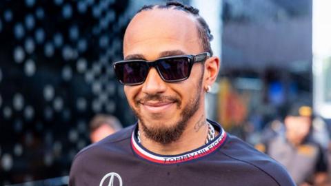 Lewis Hamilton smiles while wearing sunglasses