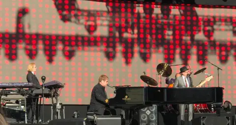 Halton Council Elton John is sitting a grand piano on stage and he is singing into a microphone. There are two musicians on either side of him playing a guitar and keyboard. There is a number of items of musical equipment on stage 