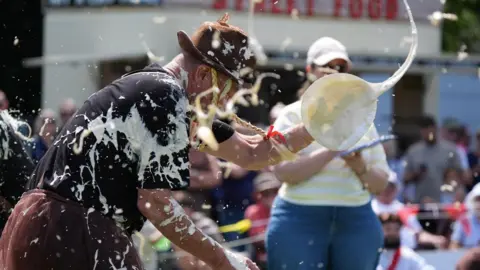 PA Media A man wearing a hat, black shirt and brown trousers is covered in custard pie. There is a woman, wearing a baseball cap, taking notes in the background. 