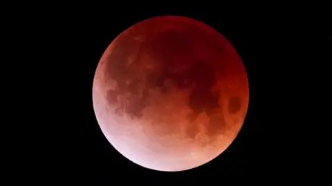 An image of the moon during a partial lunar eclipse shows it in a shade of red, caused by the lack of light hitting its surface. It is surrounded by the black openness of space.