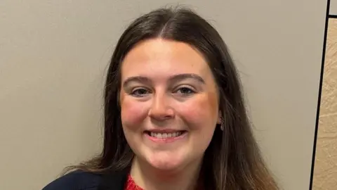 A woman with long brown hair smiling at the camera. She is wearing makeup.