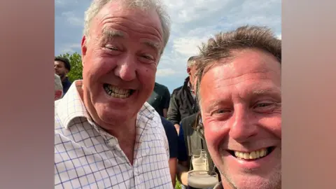 Chris Slatter A close-up selfie of Jeremy Clarkson on the left, smiling broadly and wearing a white shirt with a blue check pattern, and Chris Slatter on the right, also smiling. Chris has short brown hair. In the background other people can be seen holding drinks, their faces and bodies mostly obscured by Clarkson and Slatter's faces taking up the image