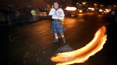 Getty Images A man in a kilt and a rugby shirt swinging a fireball in a dark street