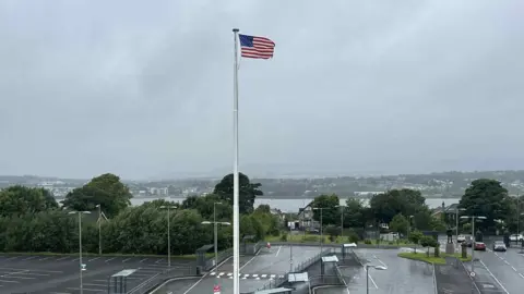 A US flag is pictured flowing in the wind on a large white flag pole.
