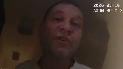 West Mercia Police A man with short hair and stubble, wearing a white T-shirt, pictured from the shoulders up in a dark room