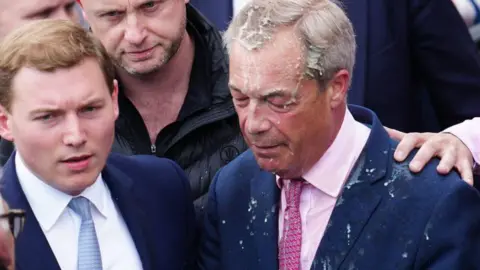 El líder del partido Reform UK, Nigel Farage, reacciona después de que una mujer le arrojara una bebida. A su izquierda se encuentra un segundo hombre, vestido con traje, camisa blanca y corbata azul.