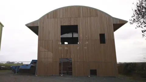 The cover of the play area is a tall structure of thin wooden planks and resembles a house. It is fenced off. There are building materials next to it.