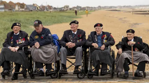 SONT Five D-Day veterans on wheelchairs on a sandy beach in Normandy. They are dressed in suits with their military badges displayed on their blazers.