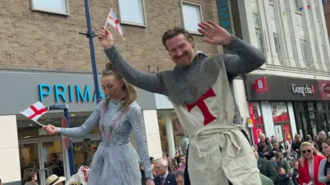 A performer dressed as St George waves while holding a St George’s flag during a parade, with another costumed performer beside them and crowds watching from the street behind.
