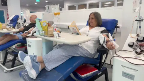 Image shows Sue sat in a blue chair with with a table in front of her. She is reeding a book. She is wearing a white top, blue striped trousers, and blue shoes. She has short hair. Her left arm is elevated on the arm rest as she is donating platelets. 