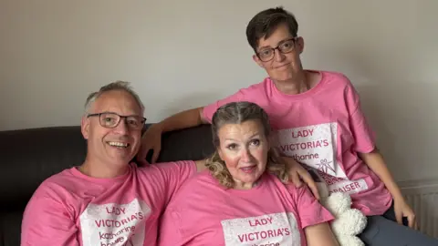 Two women and a man are sitting on a sofa, posing for a photograph. They are each wearing pink t-shirts with writing on them that reads Lady Victoria's Katharine House Fundraiser.