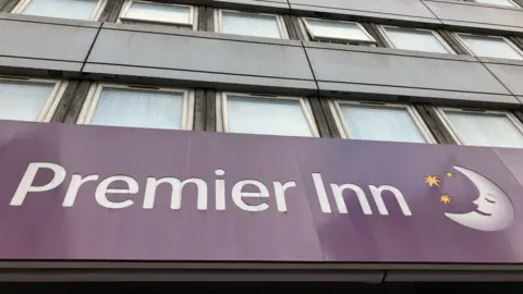 The bottom third of the picture is a sign on a Premier Inn hotel. The sign is purple with white text and the company logo is to the right of the sign, which is a crescent moon with a sleeping face on it and three stars. Above the sign two lines of hotel bedroom windows can be seen. You cannot see into the windows.