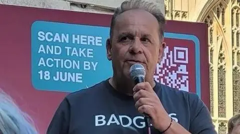 Nigel Palmer Nigel Palmer, chief executive at the Badger Trust. He has short brown hair and he is speaking to a crowd, which is out of shot, holding a microphone. He is wearing a t-shirt with the word 'badgers' on it. 