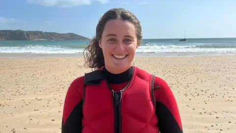 Claudia wearing a red and black wetsuit standing on an St Brelade's beach with the sea behind her. The sky is bright with light clouds, and a rocky headland curves around the bay.
