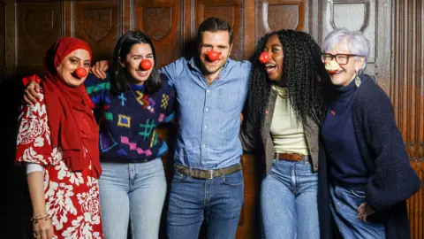 PA Media Four women and one man, all wearing red noses, standing against a wood panelled wall with their arms round each other and smiling.