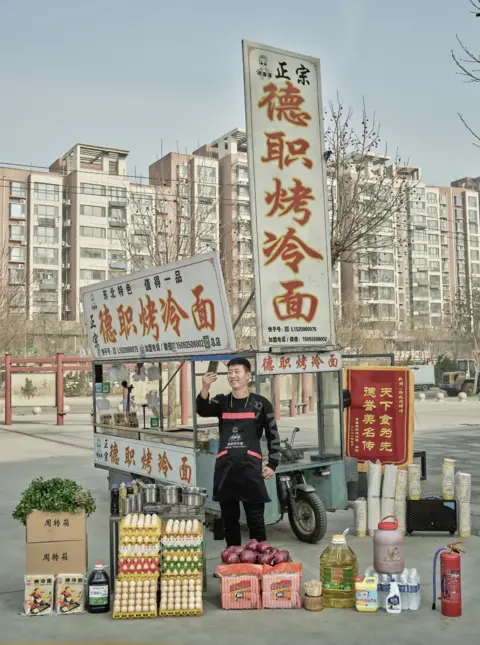 Huang Qingjun Man stands in front of a roadside hawker stall while taking a shot of himself. In front of him are trays of eggs, cooking oil, bottles of sauces and produce to cook cold grilled noodles.