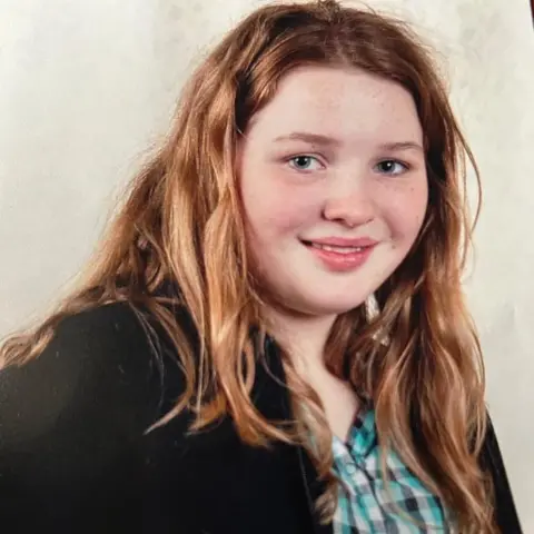 Becca Galloway A school photo-style image of Becca as a teenager, looking similar but younger, with light brown hair, a black cardigan and a green checked shirt.