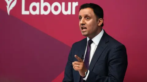 PA Media Anas Sarwar, who has short black hair, speaks while pointing his left index finger. He is wearing a dark suit and tie with a white shirt. He is in front of red Labour branding. 