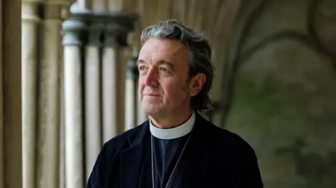 A man wearing a dog collar and robes with a chain round his neck looking into the distance. In the background are columns as found in a church or cathedral setting.