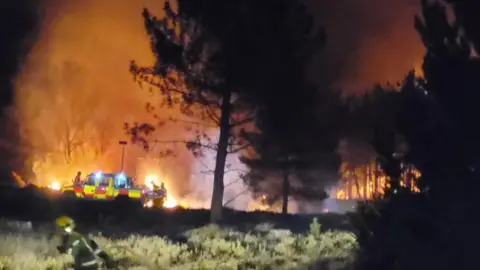 A large fire illuminates the night sky, as firefighters battle around it to get it under control.