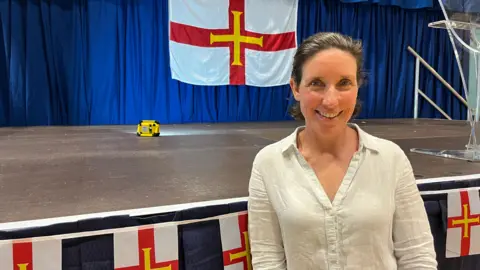 BBC A women with brown hair smiling at the camera. She is stood in front of a stage that has Guernsey flag bunting across it. There is a large Guernsey flag in the background.