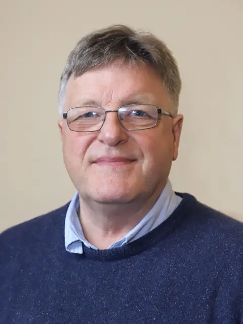 Dumfries and Galloway Council A man with grey hair and glasses in a blue jumper with a blue shirt underneath