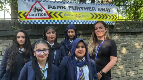 Aisha Iqbal/BBC A group of five schoolgirls in blue blazers, white shirts and striped ties stand with a woman next to some railings. Behind them is a sign that says 'park further away' and has other road safety warnings